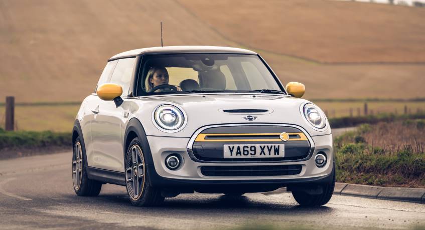 White 2 door Mini electric car with yellow wingmirrors driving on left hand side of country road in daytime