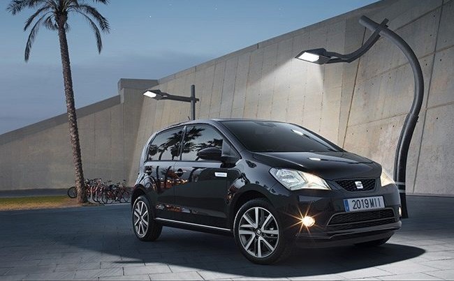 Small black 4 door Seat Mii Electric car parked under 2 street lights with its lights on at dusk