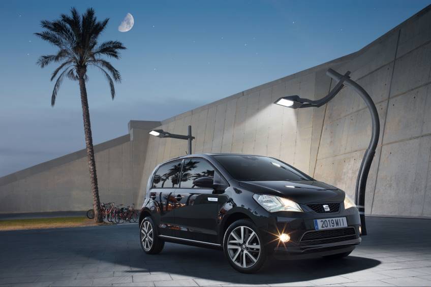 Small black 4 door Seat Mii Electric car parked under 2 street lights with its lights on at dusk