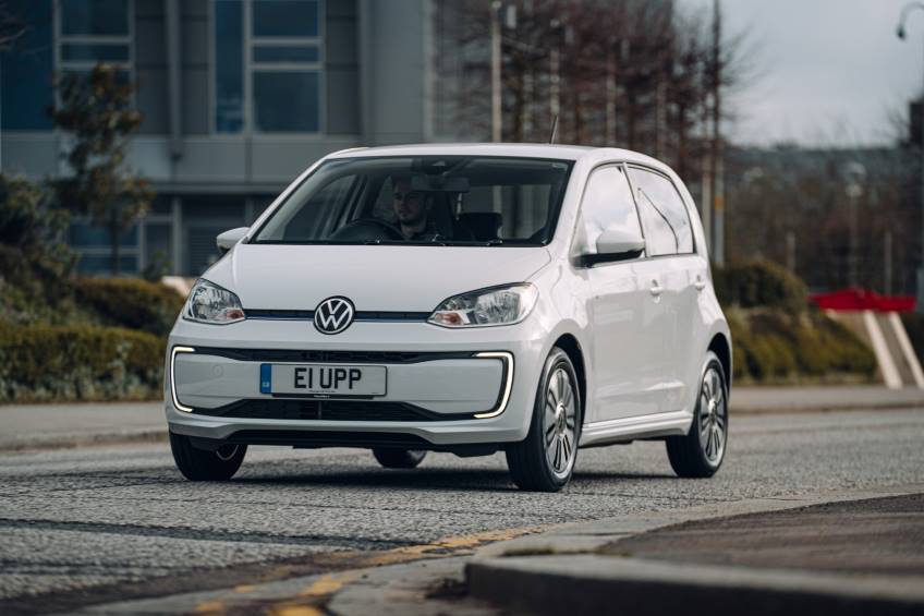 White 4 door VW Up car being driven on a road by a male with buildings in the background